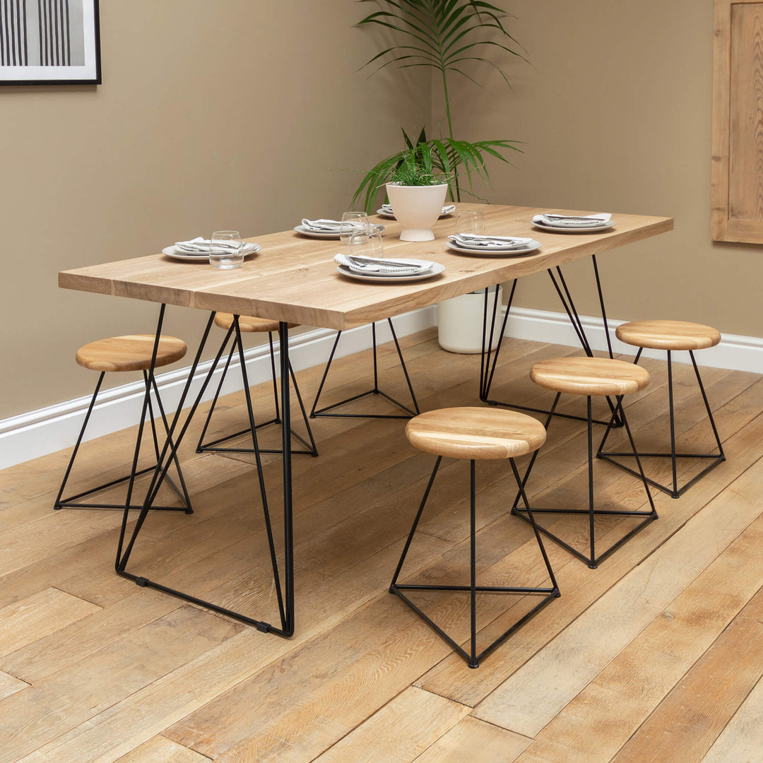 A solid oak dining table with hairpin legs made of black powder-coated steel, accompanied by wooden stools with a geometric base, set in a dining room environment.