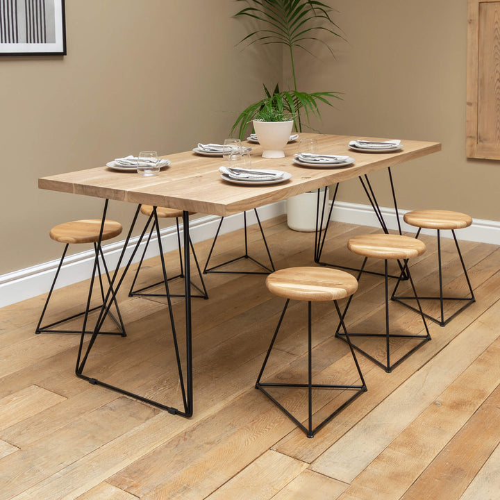 A solid oak dining table with hairpin legs made of black powder-coated steel, accompanied by wooden stools with a geometric base, set in a dining room environment.